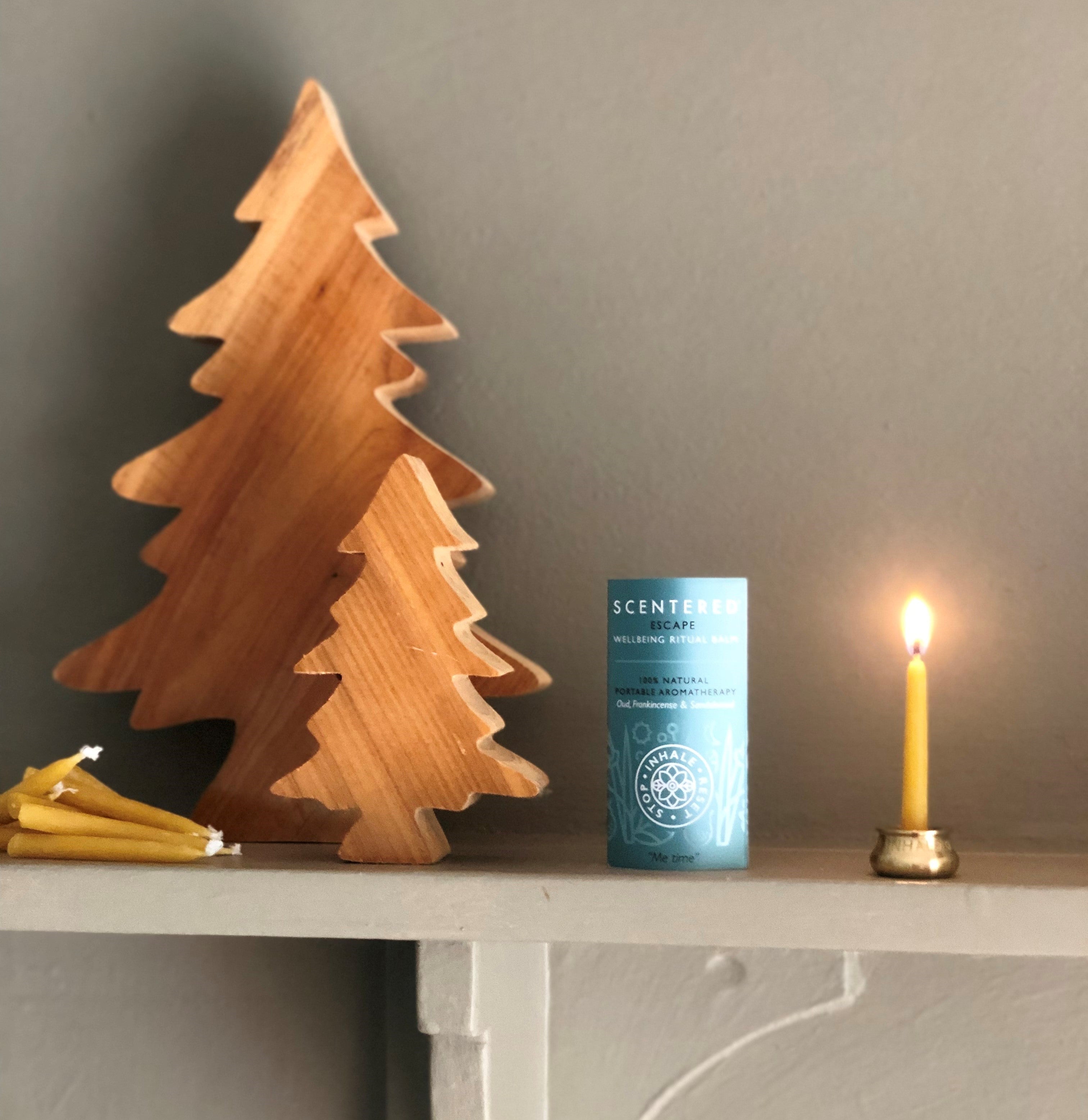 A calming holiday scene with a Scentered "Escape" Aromatherapy Balm, a lit beeswax candle, and wooden pine tree ornaments on a fireplace.