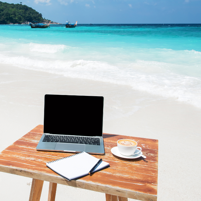 A desk and laptop, on a sandy beach by the ocean.