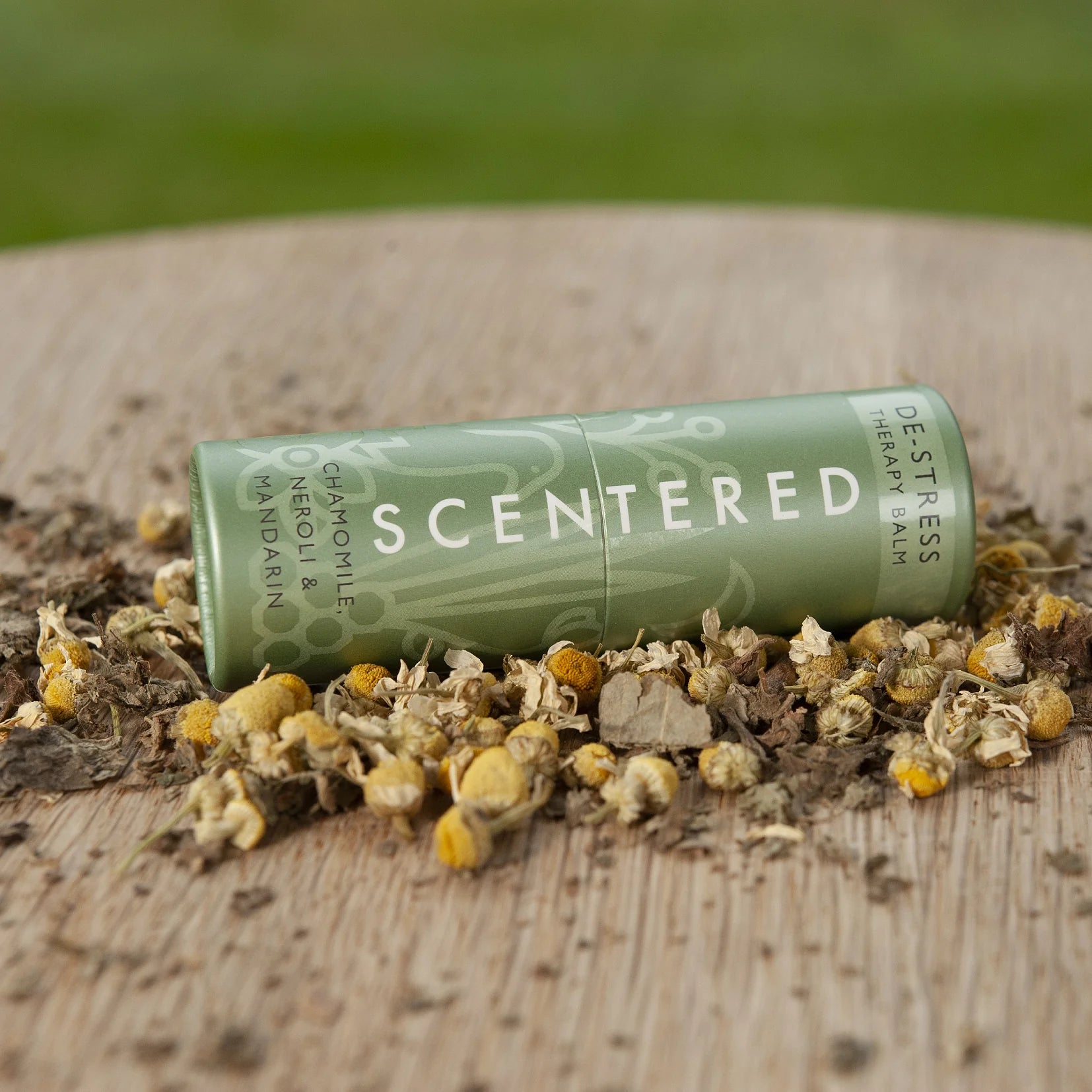 This is a picture of a Scentered De-stress Aromatherapy Balm, full size 5g, lying on its side on a table surrounded by the ingredients that are included in the balm, especially chamomile