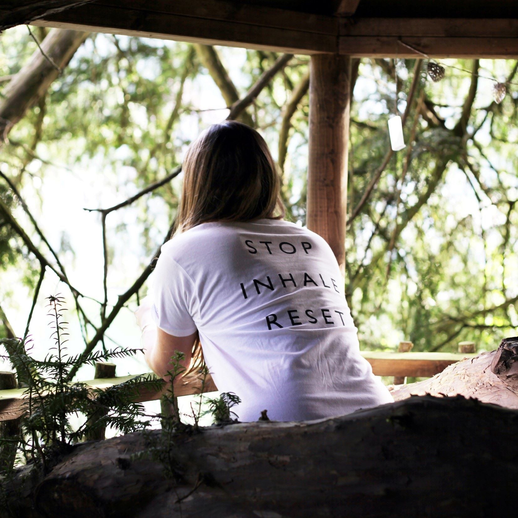 Lady in white t-shirt with Stop Inhale Reset printed on the back leaning on a wooden fence looking out onto woodlands